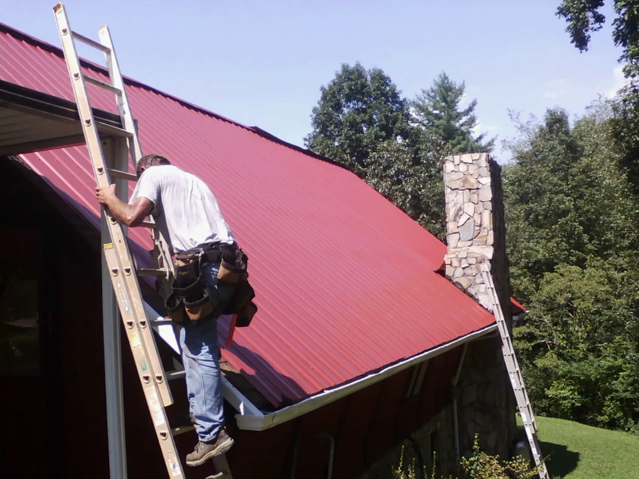 Roofer on a ladder beside a red metal roof and stone chimney — Soffit & Fascia Repair by Roofing Brothers in Wrightsville Beach